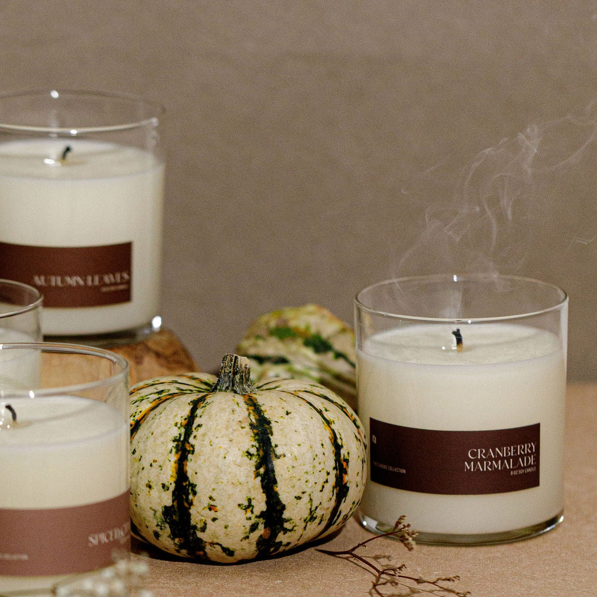A styled arrangement of several white scented candles in clear glass jars with brown labels, set on a tabletop with small gourds and a neutral wall background. Visible label text includes 'MUMBLE BAY' and 'CRANBERRY MARMOT'.