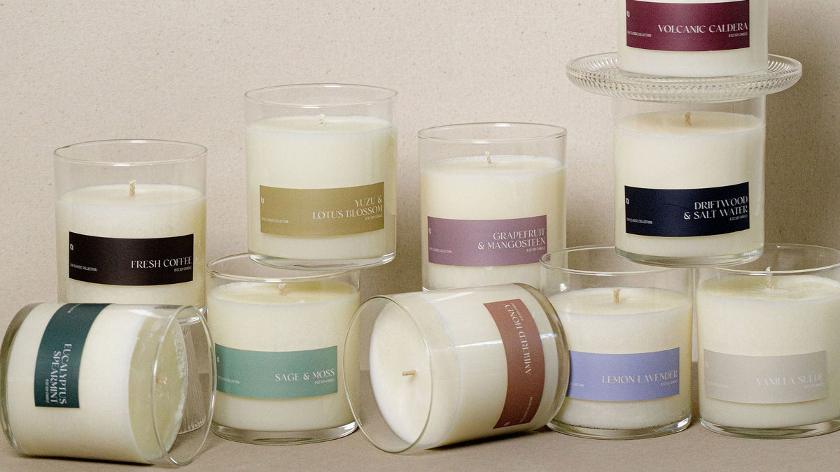 A group of white wax candles in clear glass jars arranged on a neutral beige surface against a matching textured beige background. Several candles are upright, while a few are lying down, including one on a small clear riser.